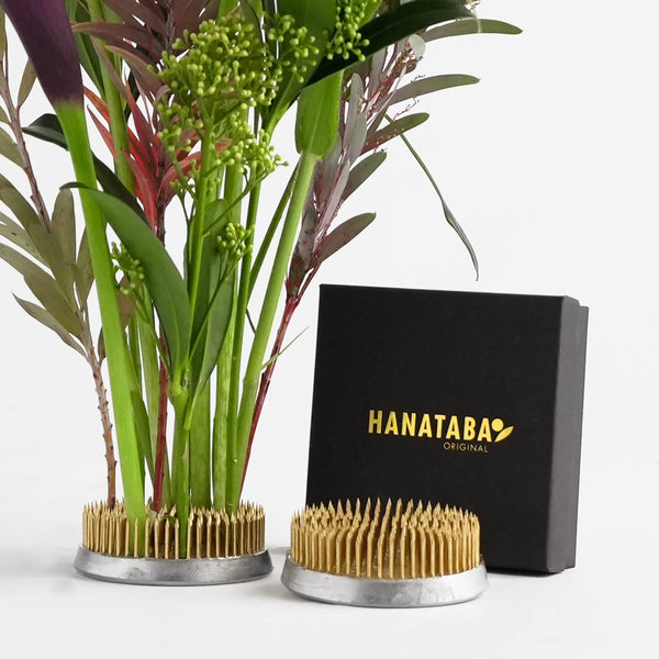 Two kenzan 70 mm flower arrangement tools and a black packaging box with "HANATABA ORIGINAL" in gold text is displayed in a bright setting. Fresh green and red stems and foliage is beautifully arranged in one of the floral frogs.