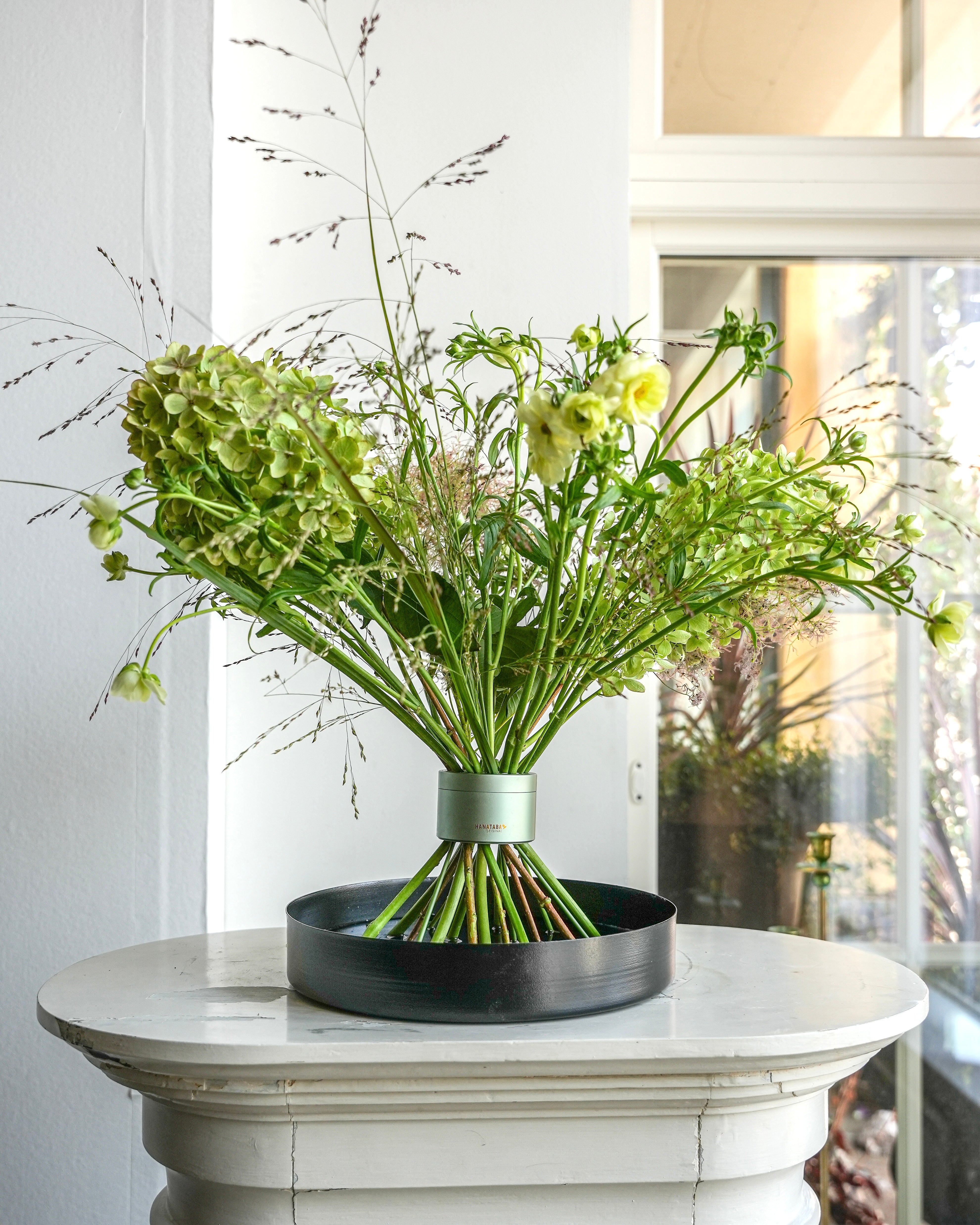 Professional bouquet with beautiful volume in Hanataba Matcha Green flower twister, displayed on a black floral tray atop a white pedestal.