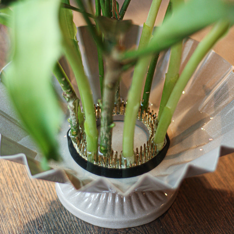 Close-up van een kenzan van 70 mm aan de basis van een bloemstuk. De koperen pinnen houden de groene stengels stevig vast boven een gegolfde glazen schaal en tonen het essentiële gereedschap in de ikebana bloemsierkunst.