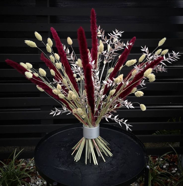 Een levendig ikebana arrangement van karmozijnrode en crèmekleurige gedroogde bloemen in een Hanataba boeket twister, tegen een zwarte achtergrond van latten.