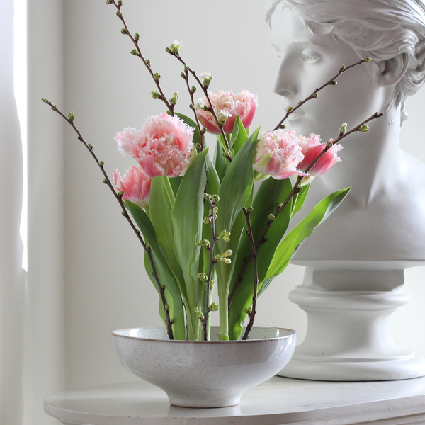 Elegant bloemstuk met roze gefranjerde tulpen en tere ontluikende takken in een gespikkelde schaal, afgewisseld met een klassiek wit borstbeeld.