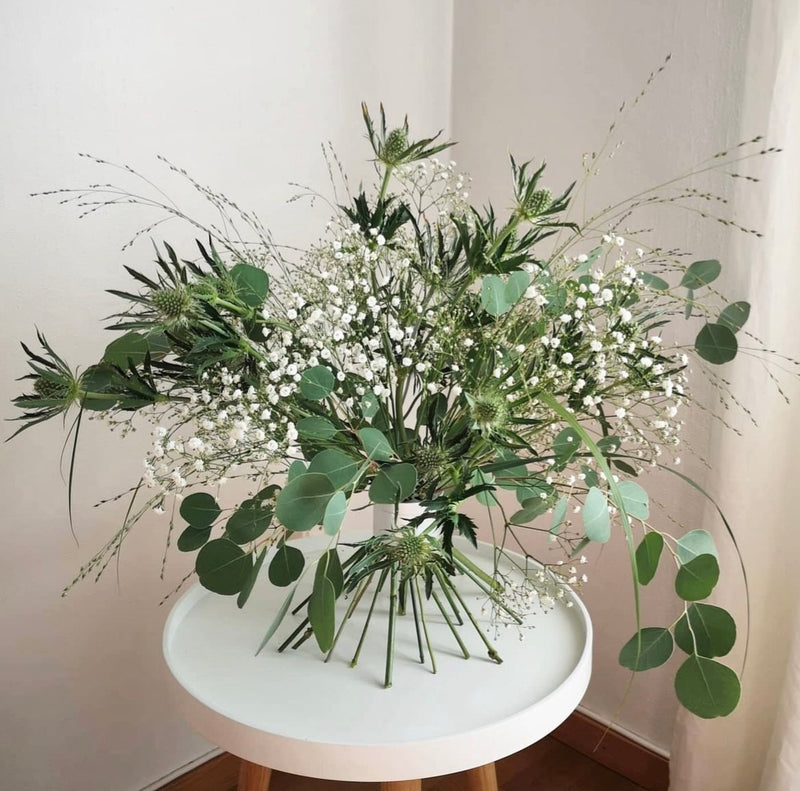 Elegant ikebana arrangement met een assortiment van groen en witte bloesems vastgezet door een Hanataba Bouquet Twister in parelmoer zilver op een witte tafel.