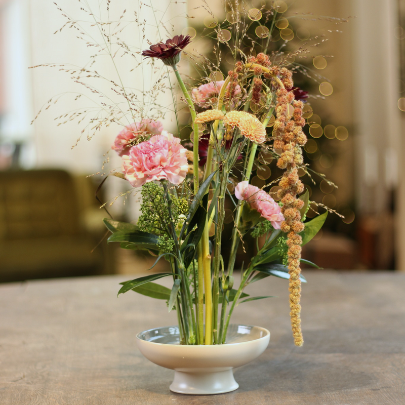 Een levendig arrangement van roze anjers, een diep bordeauxrode dahlia en delicate hangende amaranten, allemaal verankerd op een 70 mm kenzan ring, in een ondiepe witte schaal op een houten tafel, die een gevoel van harmonie en natuurlijke elegantie oproept.