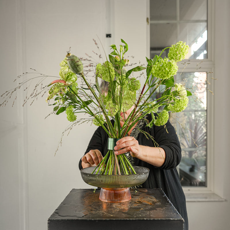 Een bloemist maakt een professioneel bloemstuk met viburnum snijbloemen in Hanataba Ivy Green, uitgestald in een gestreepte ronde glazen schaal.