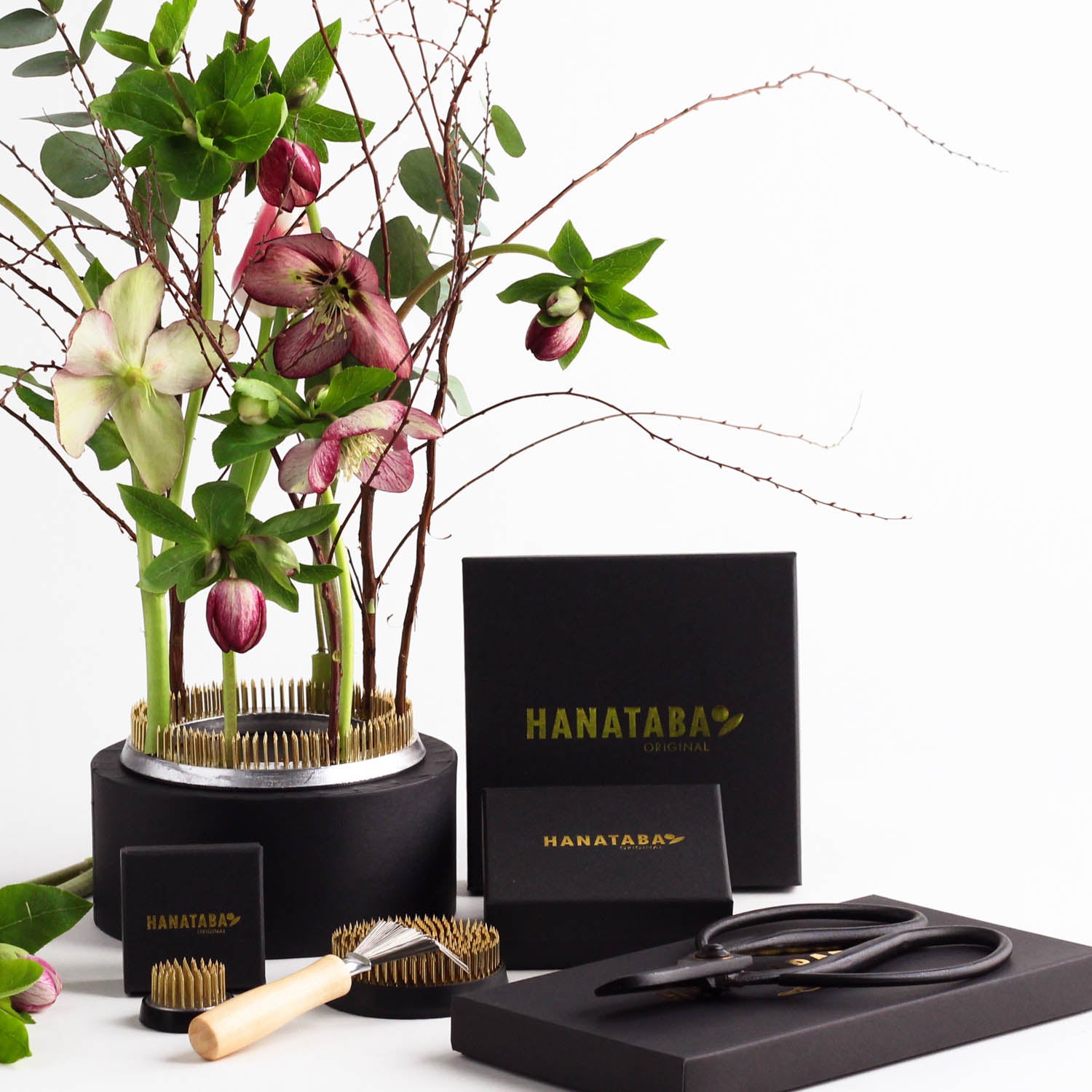 Hanataba's Flower Frog Trio & Tool Set is elegant gerangschikt met levendige roze bloemen en groene bladeren in een ronde 120 mm kenzan ring. De set bestaat uit een 70 mm en 34 mm bloemenkikker, gecombineerd met een zwarte Hanataba verpakking, een schoonmaakborstel en een Japanse tuinschaar voor professioneel bloemwerk.