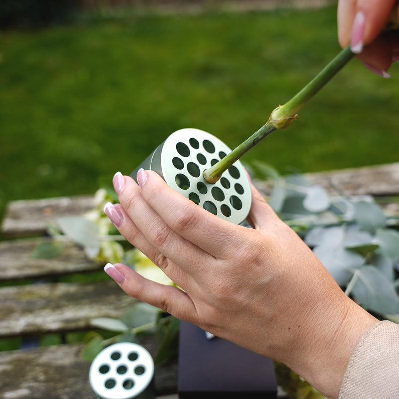 Een persoon met roze nagellak schikt eucalyptus en bloemen door een steel in een Hanataba Matcha Green boeket twister tool te steken boven een houten tafel in de tuin.