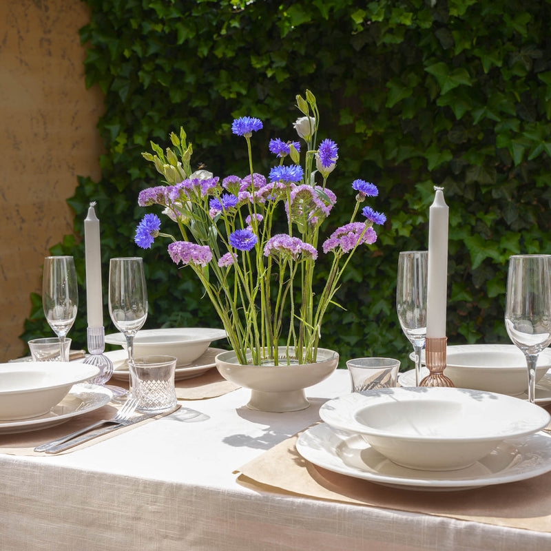 Elegant outdoor table setting with white dishes, glassware, tall candles, and a centerpiece of purple and blue flowers arranged in a flower frog ring from Hanataba. The tablecloth is white, and the backdrop features lush green ivy.