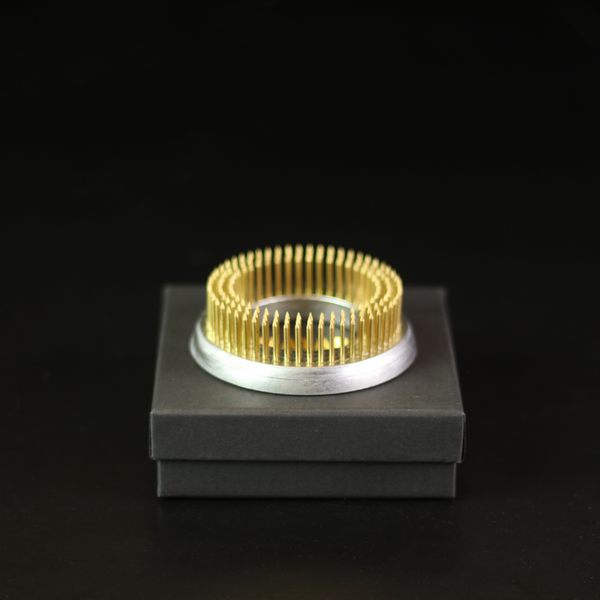 Een 70 mm kenzan ring met gouden pinnen, ontworpen voor bloemstukken, weergegeven op een donkergrijze doos tegen een zwarte achtergrond, die het glanzende metaal en de precisie benadrukt.