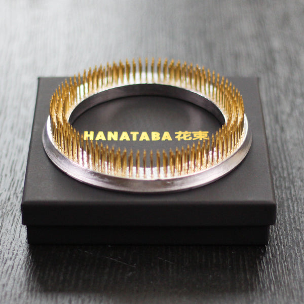 Een gouden Hanataba kenzan ring met een zilveren basis, gepresenteerd op een zwarte displaydoos met gouden tekst, met elegant Japans bloemkunstgereedschap.