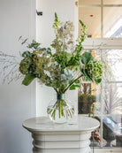 A clear glass vase with an arrangement of green and white flowers, including hydrangeas and eucalyptus, sits on a small white round table. The Hanataba Ivy Green bouquet stands out as sunlight streams through a tall window in the background.