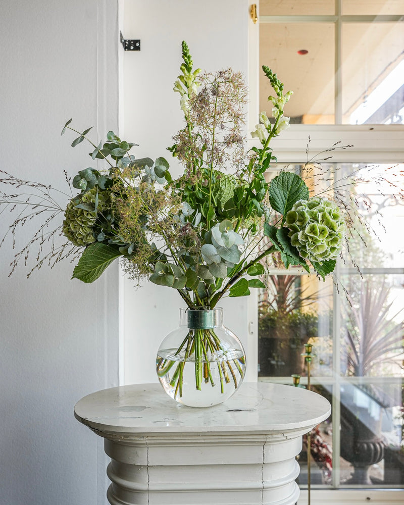 Een heldere glazen vaas met een arrangement van groene en witte bloemen, waaronder hortensia's en eucalyptus, staat op een kleine witte ronde tafel. Het Hanataba Ivy Green boeket valt op terwijl het zonlicht door een hoog raam op de achtergrond schijnt.