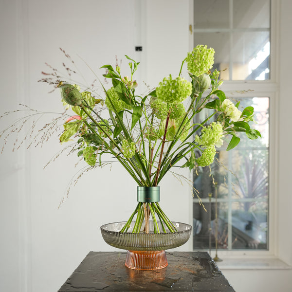 Prachtig boeket van gras- en viburnumbloemen, gerangschikt in een Hanataba Ivy Green gereedschap, uitgestald in een glazen schaal met water op een zwarte tafel bij een raam in een helder witte kamer.