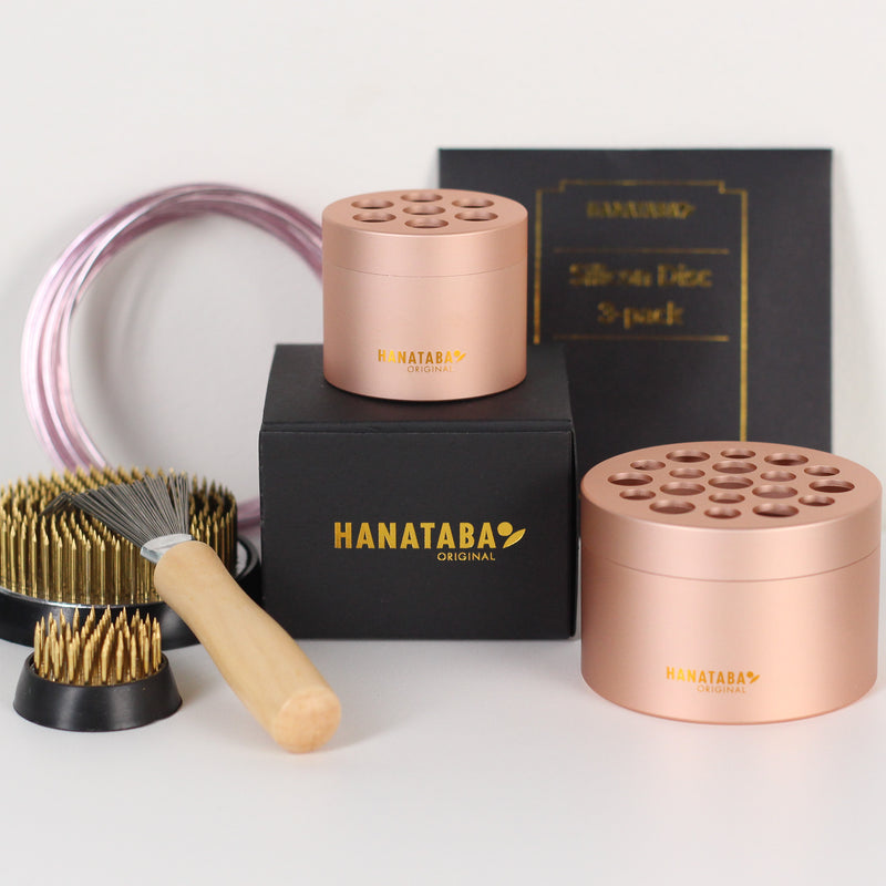 Een set Hanataba Original bloemschikgereedschap, bestaande uit twee Hanataba Pink Champagne bloemenhouders, een grotere kenzan (70mm), een kleinere kenzan (34mm) en een kenzan harkkwast met houten steel. De items zijn gerangschikt op een zwarte doos met gouden Hanataba logo belettering.