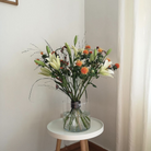 Minimalistisch bloemstuk in Ikebana-stijl met witte lelies en oranje Carthamus in een heldere vaas met een Hanataba Spiral Stem Holder op een witte tafel.