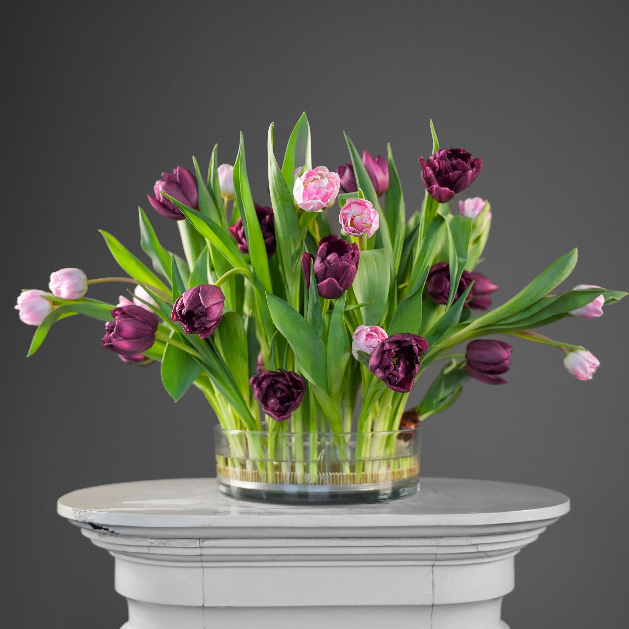 Lush bouquet of deep purple and pink tulips artistically arranged with the support of a 200mm Kenzan Fakir ring, displayed on a classic white pedestal.