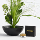 A minimalist flower frog floral arrangement with green plants elegantly sits in a black bowl, accompanied by a small black box labeled "HANATABA ORIGINAL" with a kenzan 34 mm in front of it. The scene displays crisp elegance against a plain white background.