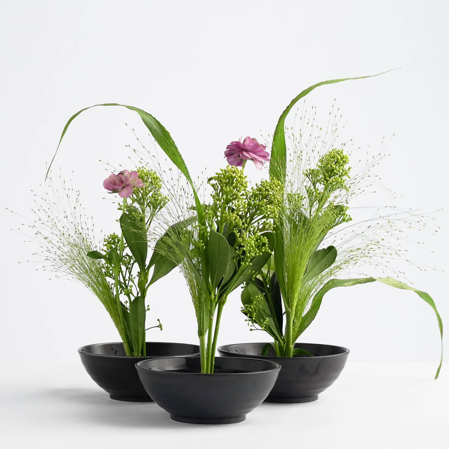 Three black bowls, each showcasing a flower bouquet of green foliage, tall grasses, and pink flowers securely placed in flower frogs, serve as elegant table decor against a white background. The minimalist design beautifully emphasizes natural beauty and simplicity.