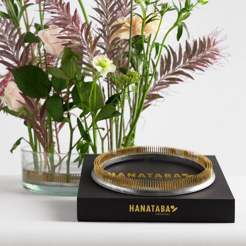 A kenzan floral arrangement with delicate pink and white flowers and green leaves in a glass vase, accentuated by a 200 mm flower frog ring that ensures perfect placement. In the foreground, there's a Hanataba Original branded packaging box displaying a kenzan ring with golden spikes on top of the box. 