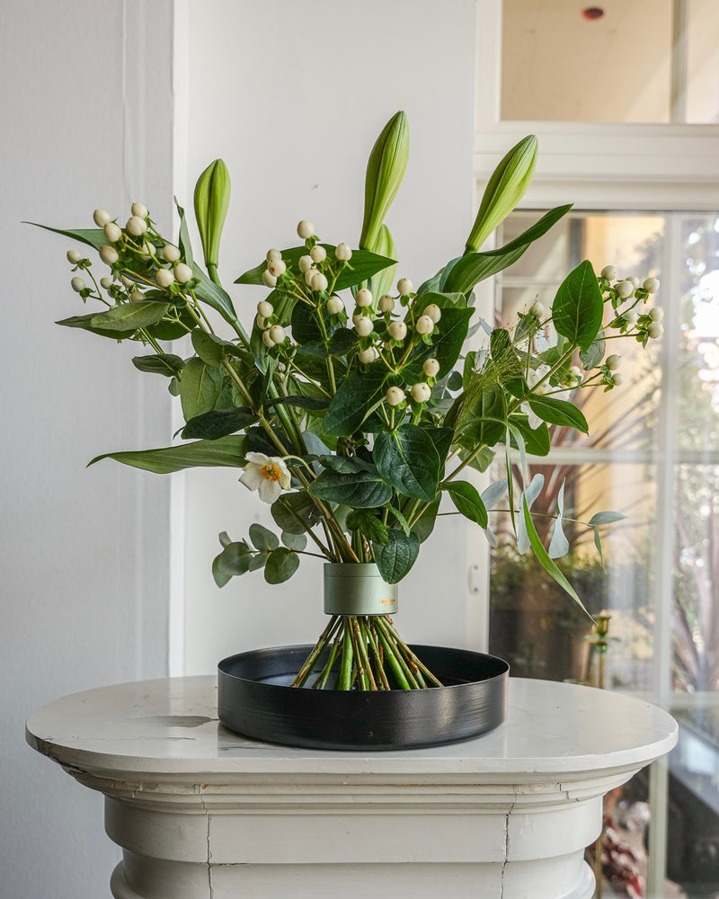 Prachtig bloemstuk met lelies op een zwarte bloementray op een witte sokkel in een mooie witte kamer met ramen met panelen.