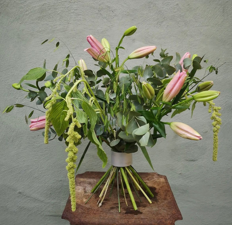 Een weelderig bloemstuk met roze lelies en groene accenten, bijeengehouden door een Hanataba Bouquet Twister in parelmoer zilver, tegen een grijze muur met structuur.