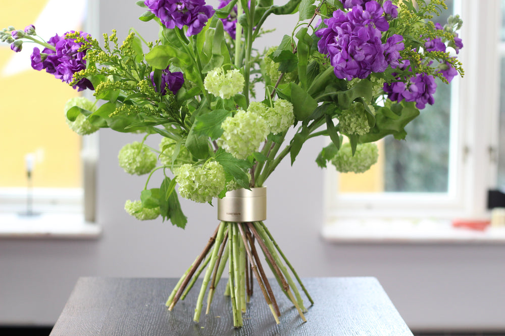 Levendig boeket met paarse stengels en groene viburnum gearrangeerd met een Hanataba Original Spiral Stem Holder in champagne goud op een tafel bij het raam.