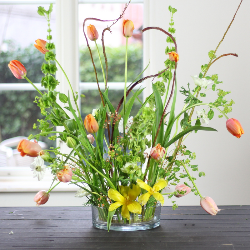 Een levendig arrangement van tulpen en gemengd groen rechtop in een 200mm kenzan ring, geplaatst op een donkere houten tafel bij een raam.