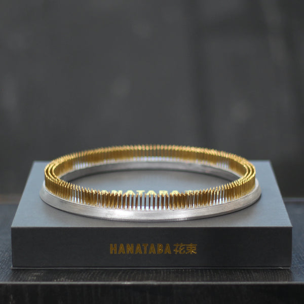 Een glanzende 200 mm Kenzan ring rust op een slanke grijze presentatiedoos met de gouden inscriptie "HANATABA 花束," een toonbeeld van elegantie en functionaliteit in bloemschikken.