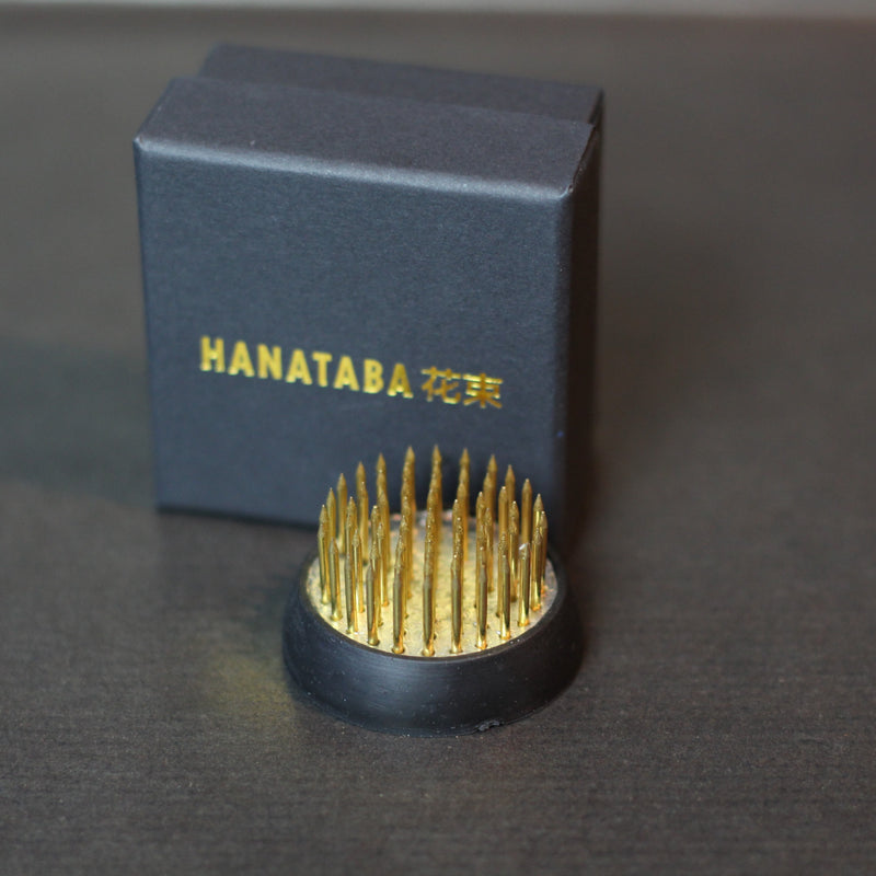 Een 34 mm Kenzan ring van Hanataba met gouden spikes, uitgestald naast zijn donkerblauwe doosje met gouden letters.