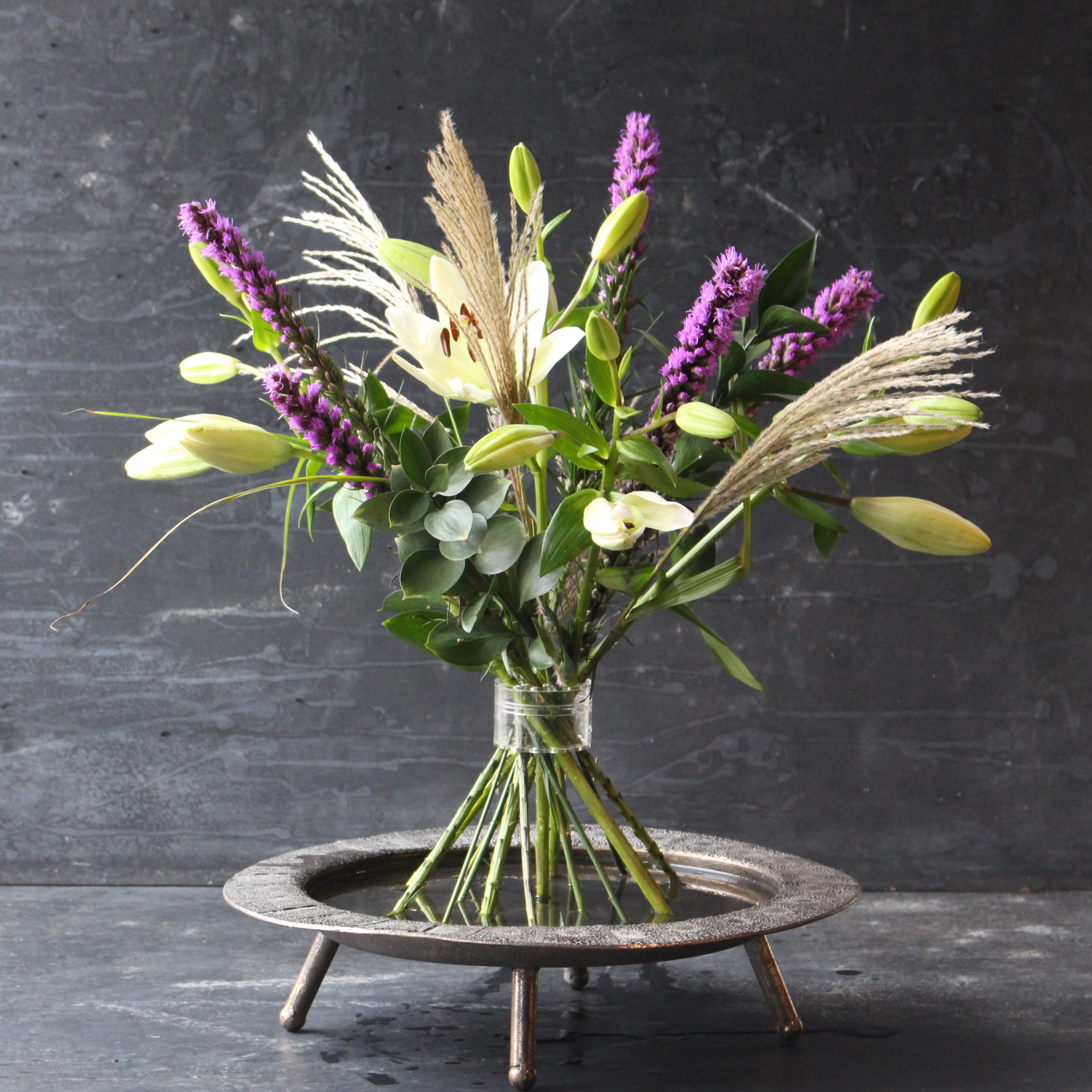 Een Hanataba Bouquet Twister Crystal Clear met weelderig groen, paarse bloemen en delicaat gras, gerangschikt in een boeket twister op een donkere achtergrond.