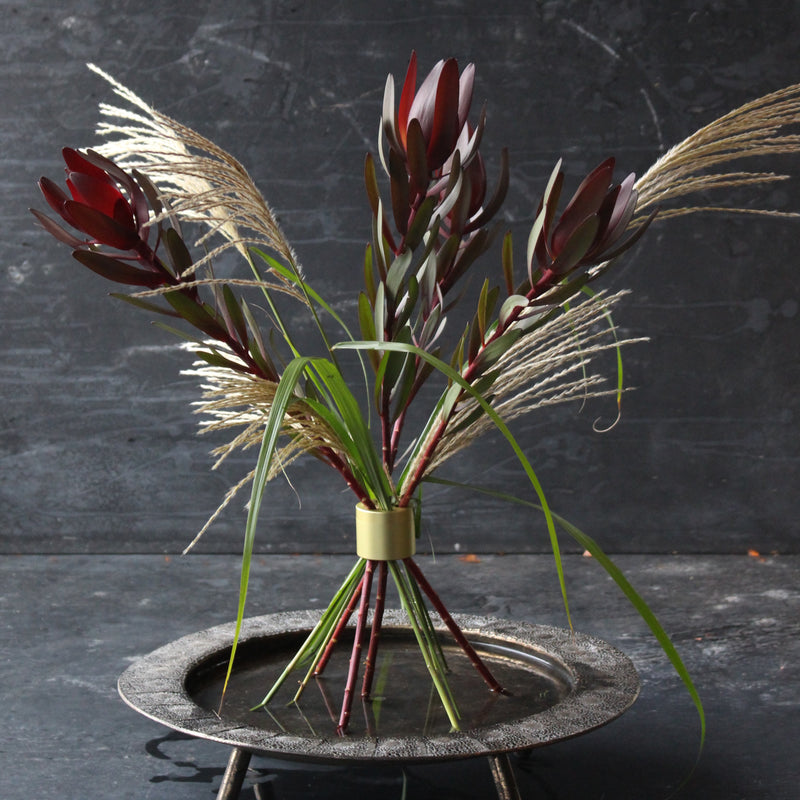 Een smaakvol bloemstuk met diep bordeauxrode leucadendronbloemen en delicate wilde grassen, elegant bijeengehouden door een Hanataba in champagnegoud.