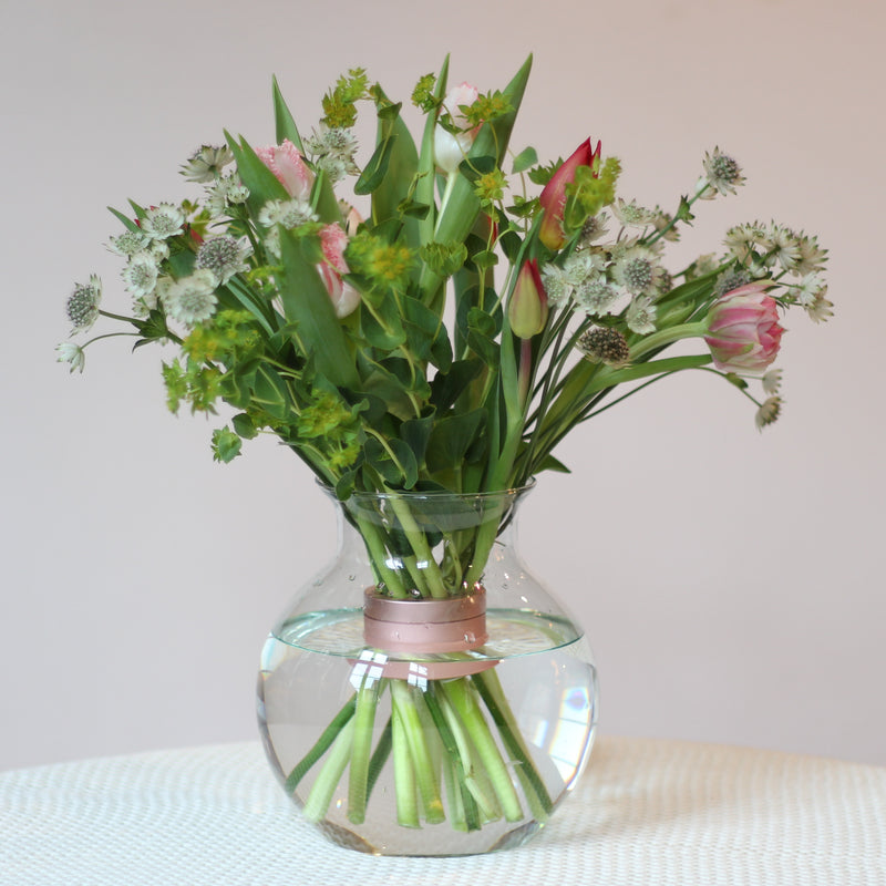 Een vers boeket van tulpen en diverse soorten groen, vastgezet door een Pearly Pink Champagne Hanataba boeket twister, staat in een heldere, ronde vaas en biedt een mix van elegantie en lente charme.