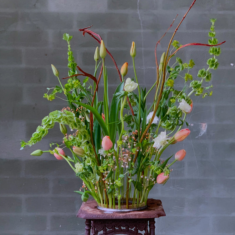 Een levendig boeket met tulpen en veel groen, zorgvuldig geschikt met een 200 mm Kenzan ring op een sierlijke houten sokkel tegen een grijze bakstenen muur.