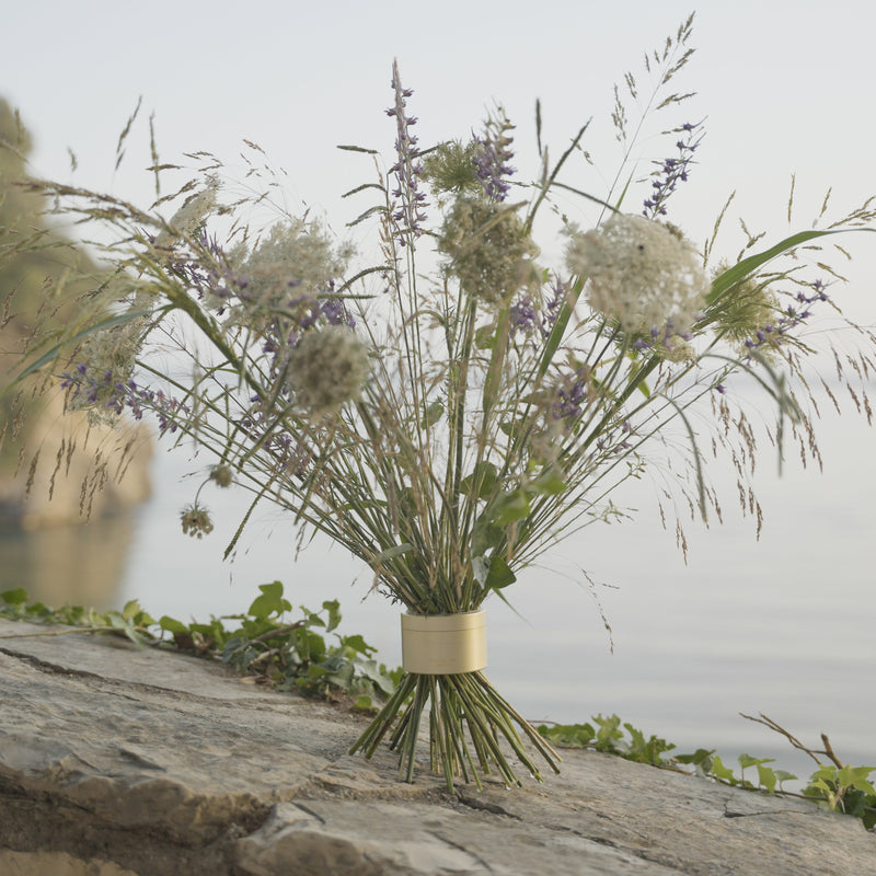 Wilde bloemen, waaronder paarse aren en witte trossen, kwamen samen in een Champagne Gold Hanataba boeket twister op een rotsachtige richel met uitzicht op kalm water.