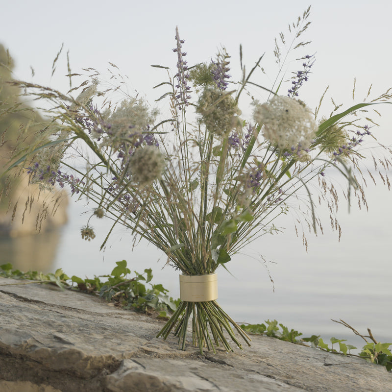 Wilde bloemen en grassen in een Hanataba Bouquet Twister Champagne Gold, geplaatst op een rots met sereen water op de achtergrond, wat natuurlijke rust uitstraalt.